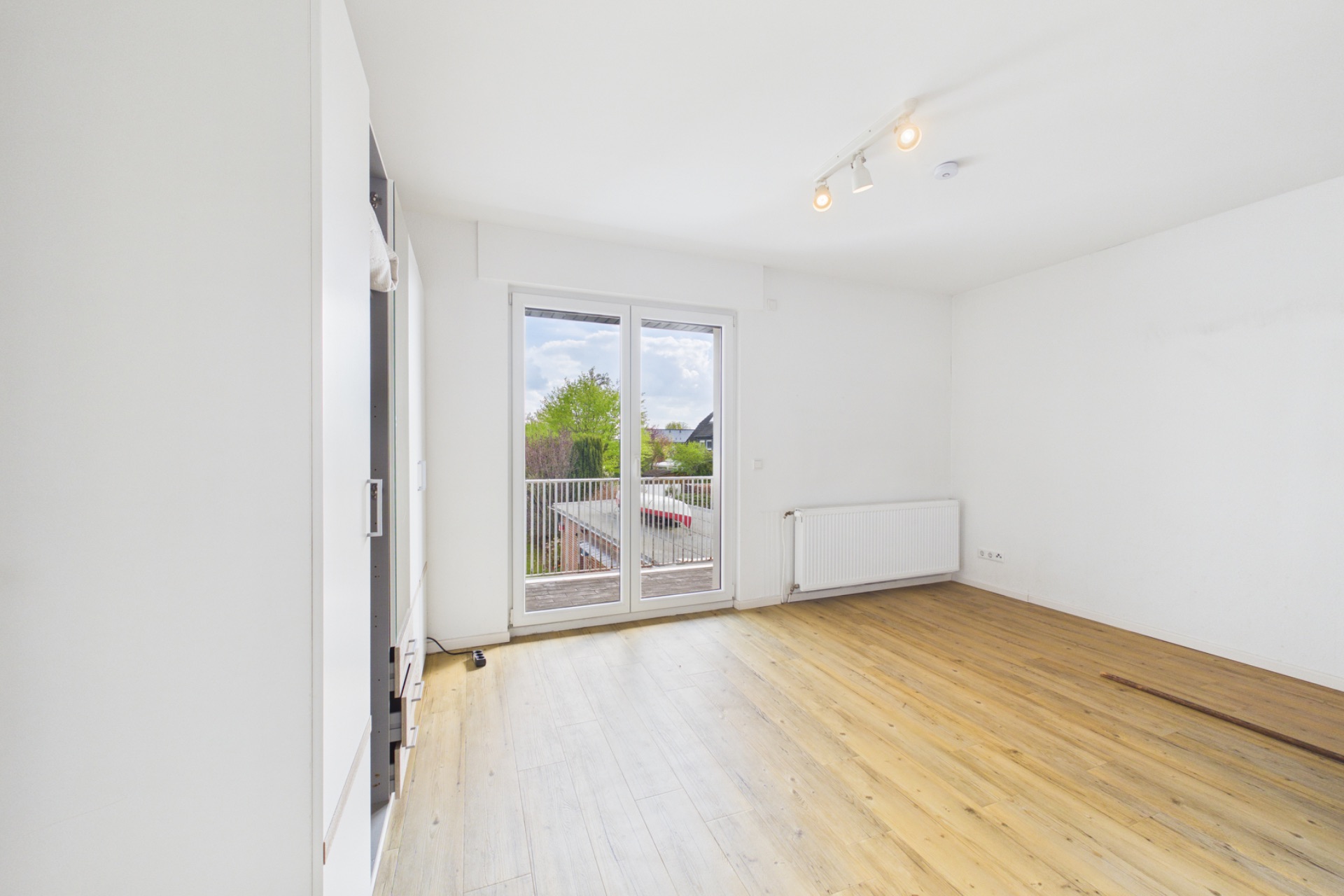 Heller Raum mit Holzboden, Doppeltür zum Balkon und Heizkörper an der Wand.