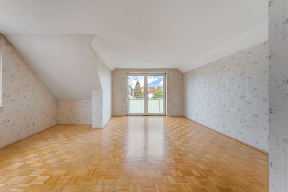 Großer, leerer Raum mit Parkettboden, gemusterter Tapete und bodentiefem Fenster mit Balkon.
