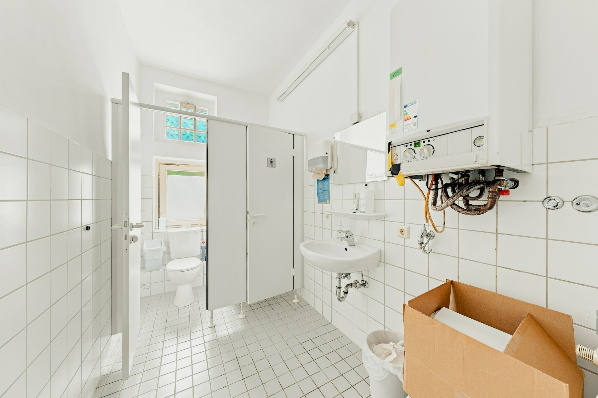 Heller Raum mit WC-Kabinen, Waschbecken, Heizung und Karton auf Fliesenboden.