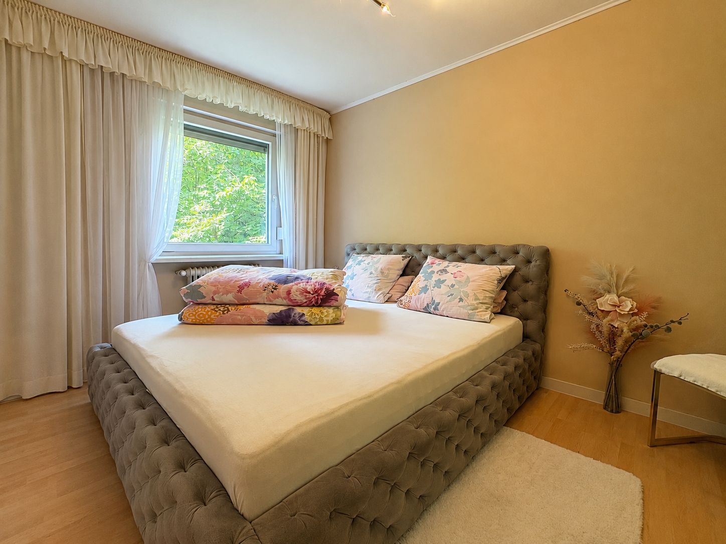 Schlafzimmer mit großem Bett, Blumenkissen, Fenster mit Vorhängen und Dekovase auf Holzfußboden.