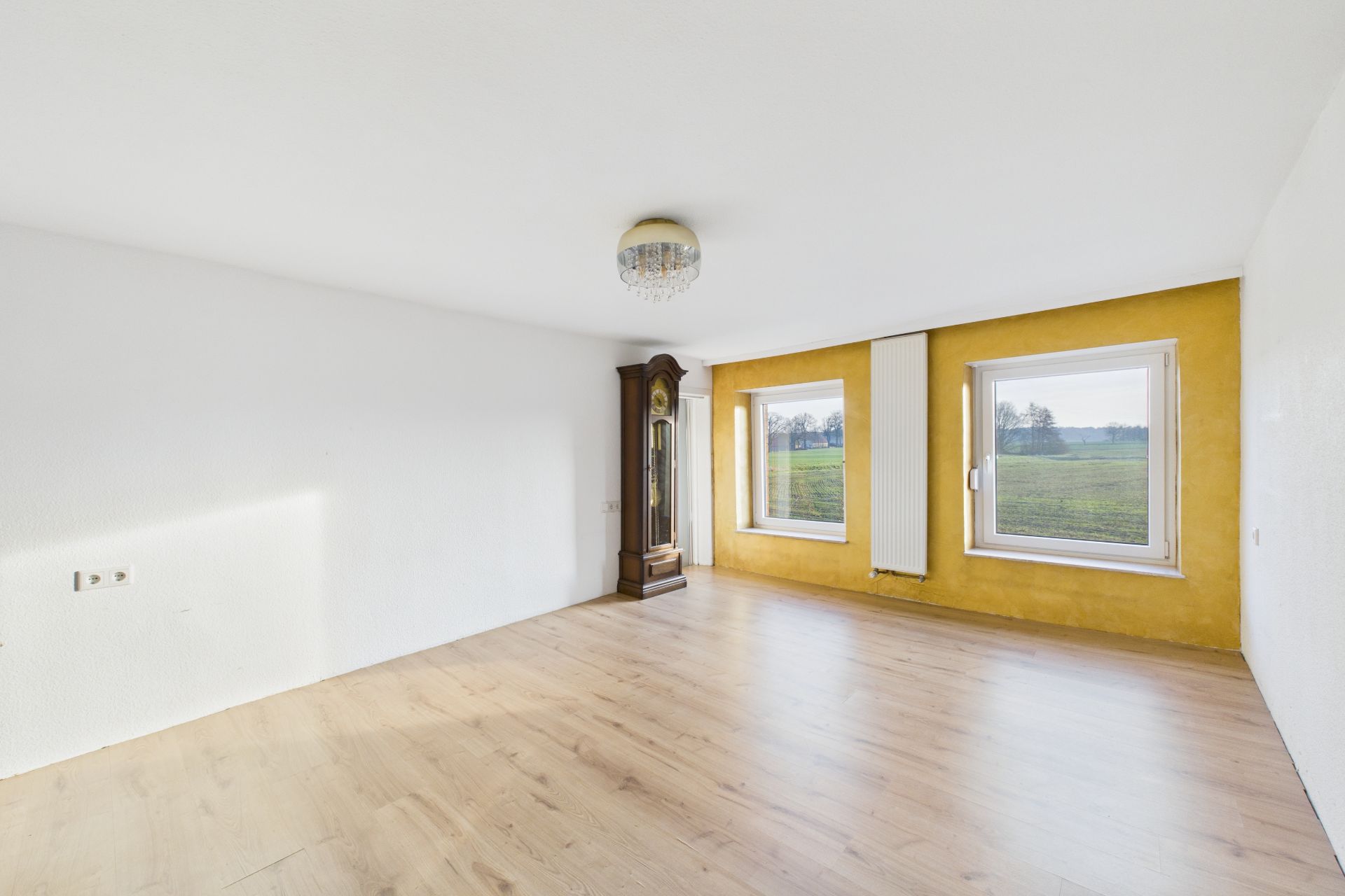 Helles Zimmer mit Holzfußboden, Standuhr, zwei Fenstern und gelber Wand.