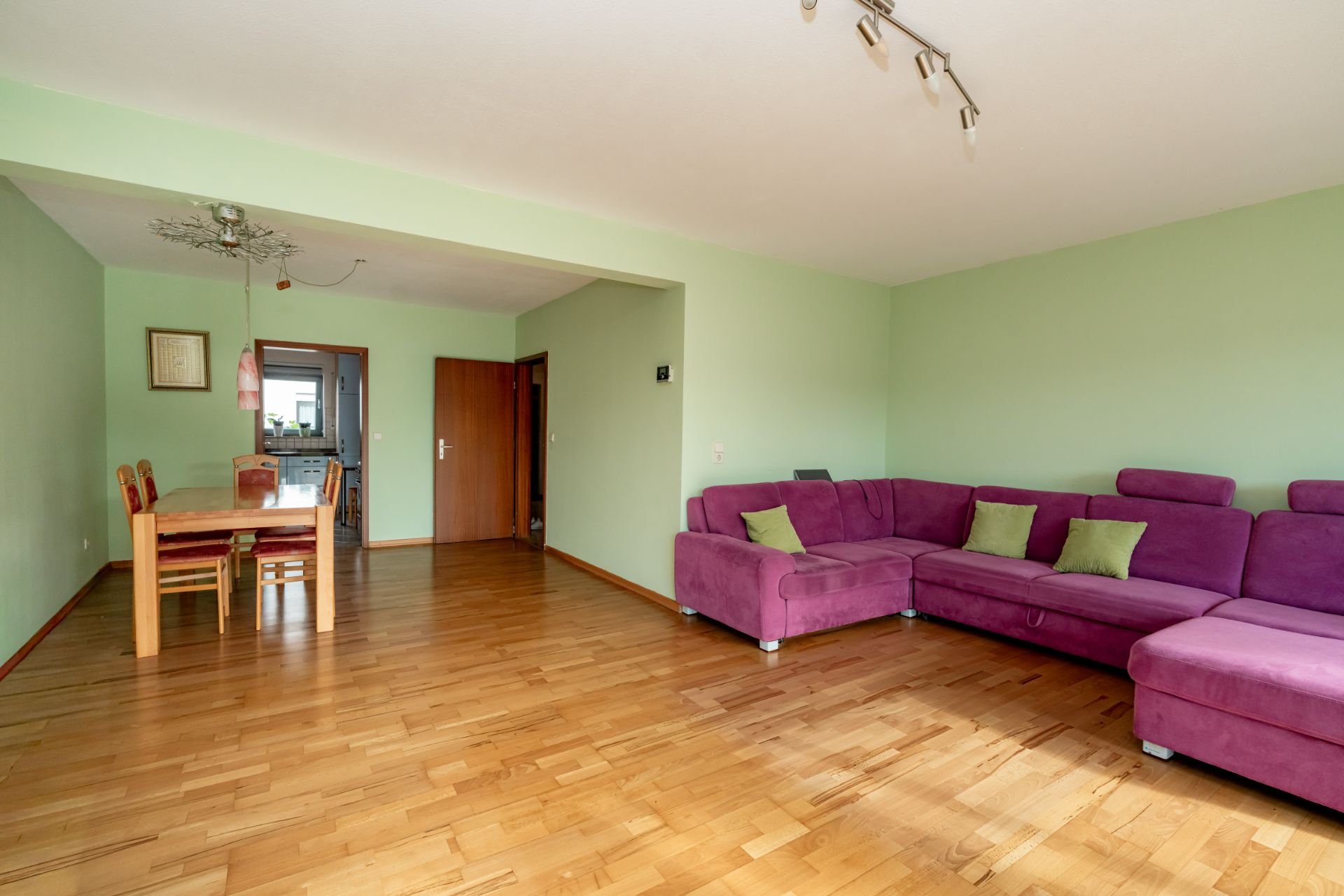 Wohnzimmer mit lila Ecksofa, Holzboden und Essbereich mit Tisch und Stühlen.