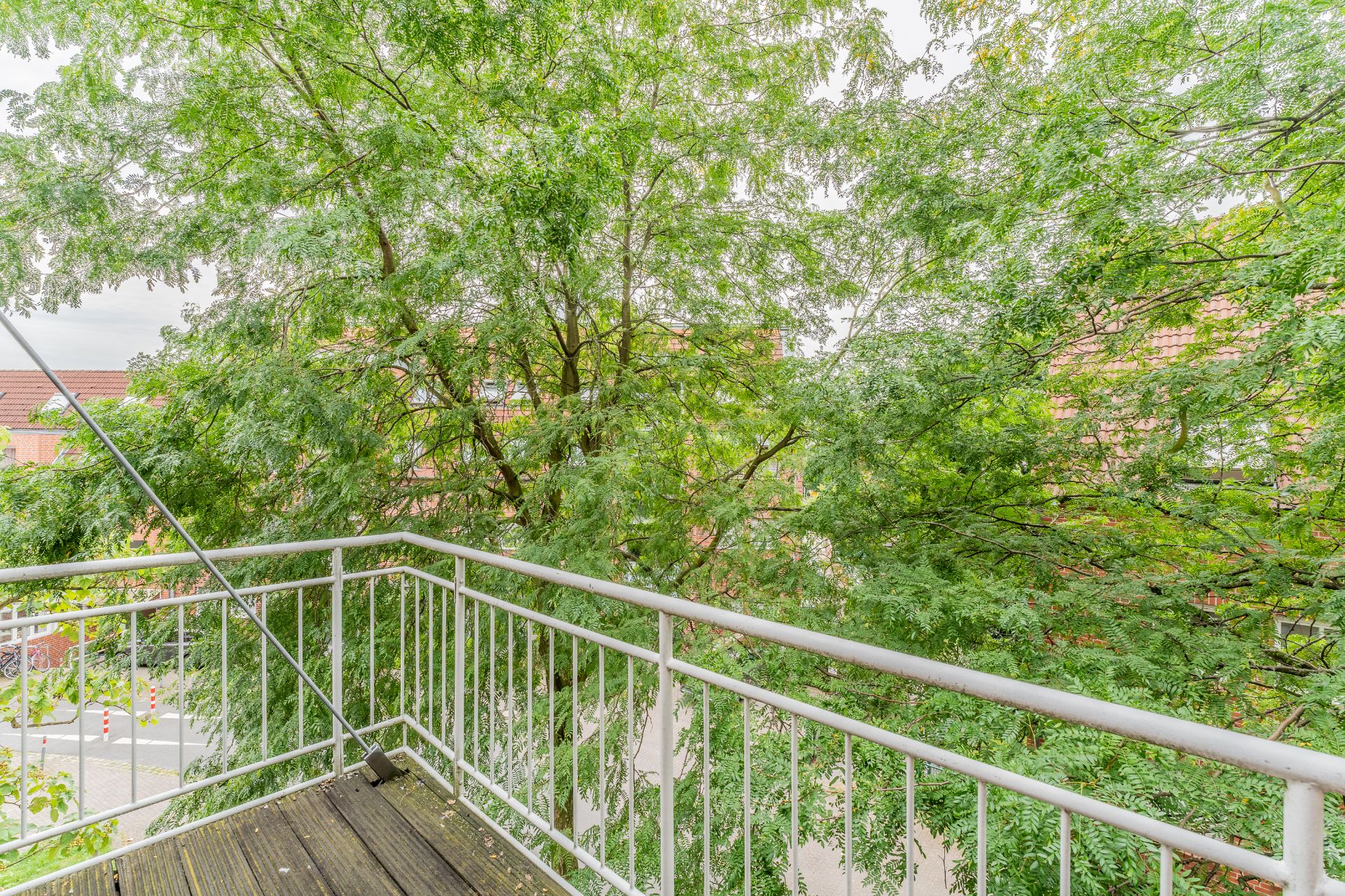 Balkon mit Metallgeländer, umgeben von grünen Bäumen und Blick auf Ziegeldächer.