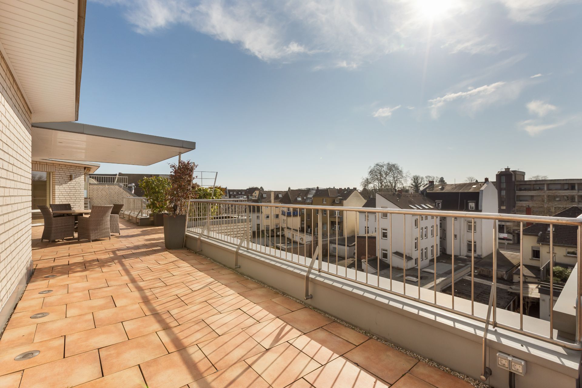 Große Terrasse mit Tisch, Stühlen und Pflanzen, Blick auf Wohnhäuser bei sonnigem Wetter.