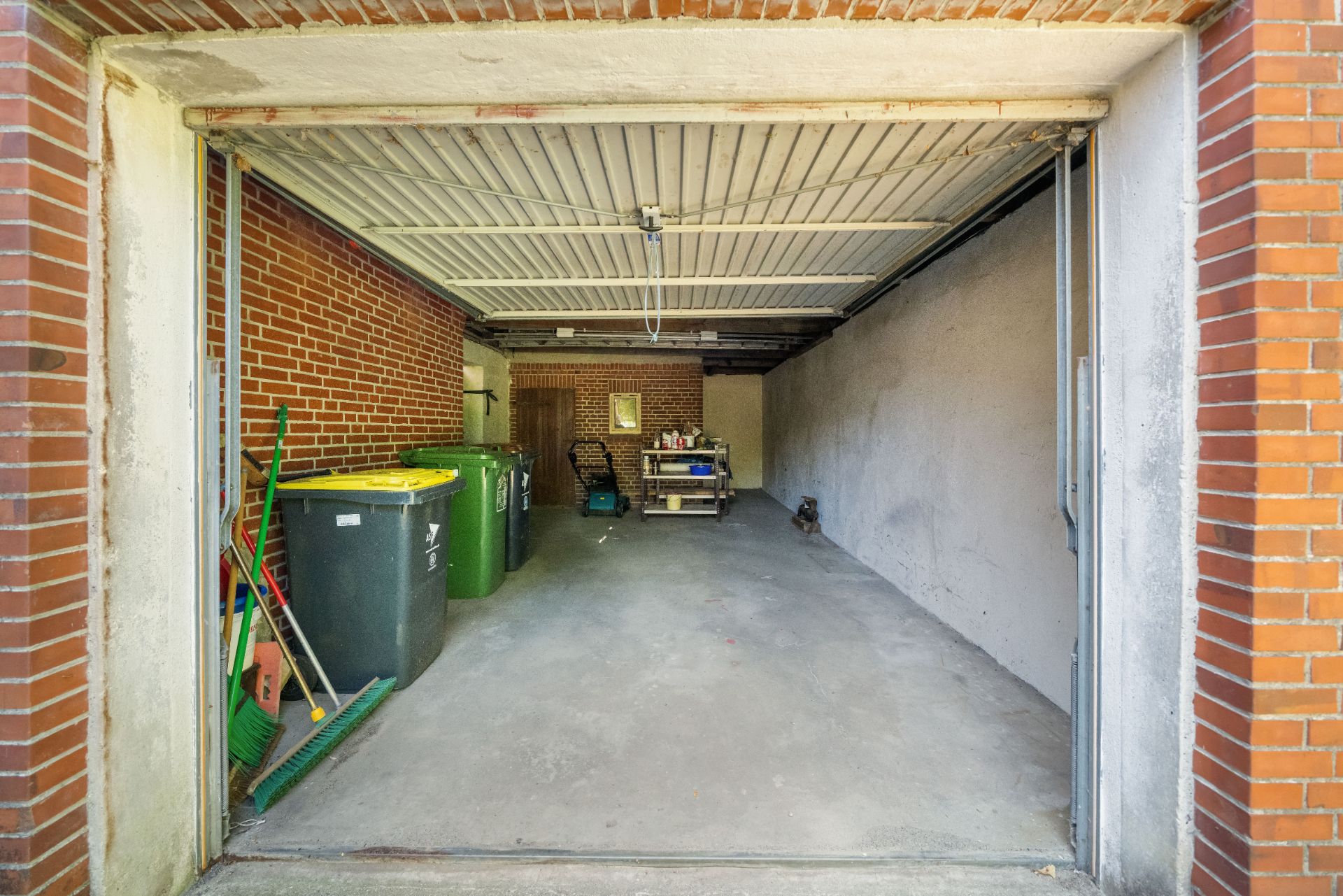 Offene Garage mit Mülltonnen und Regalen an den Wänden, Boden aus Beton.