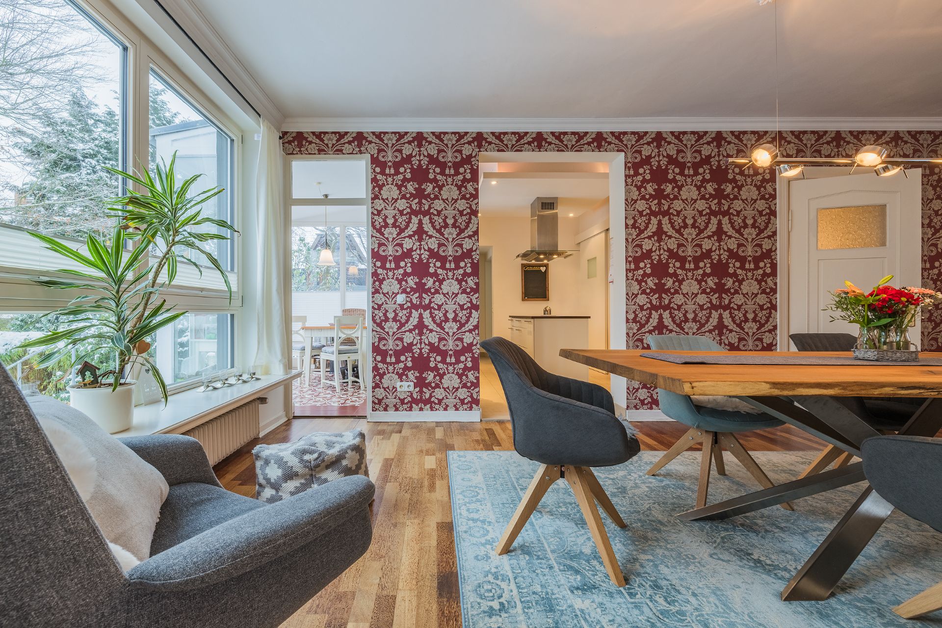Esszimmer mit Holztisch, blauen Stühlen, roter Tapete und großen Fenstern mit Pflanzen.