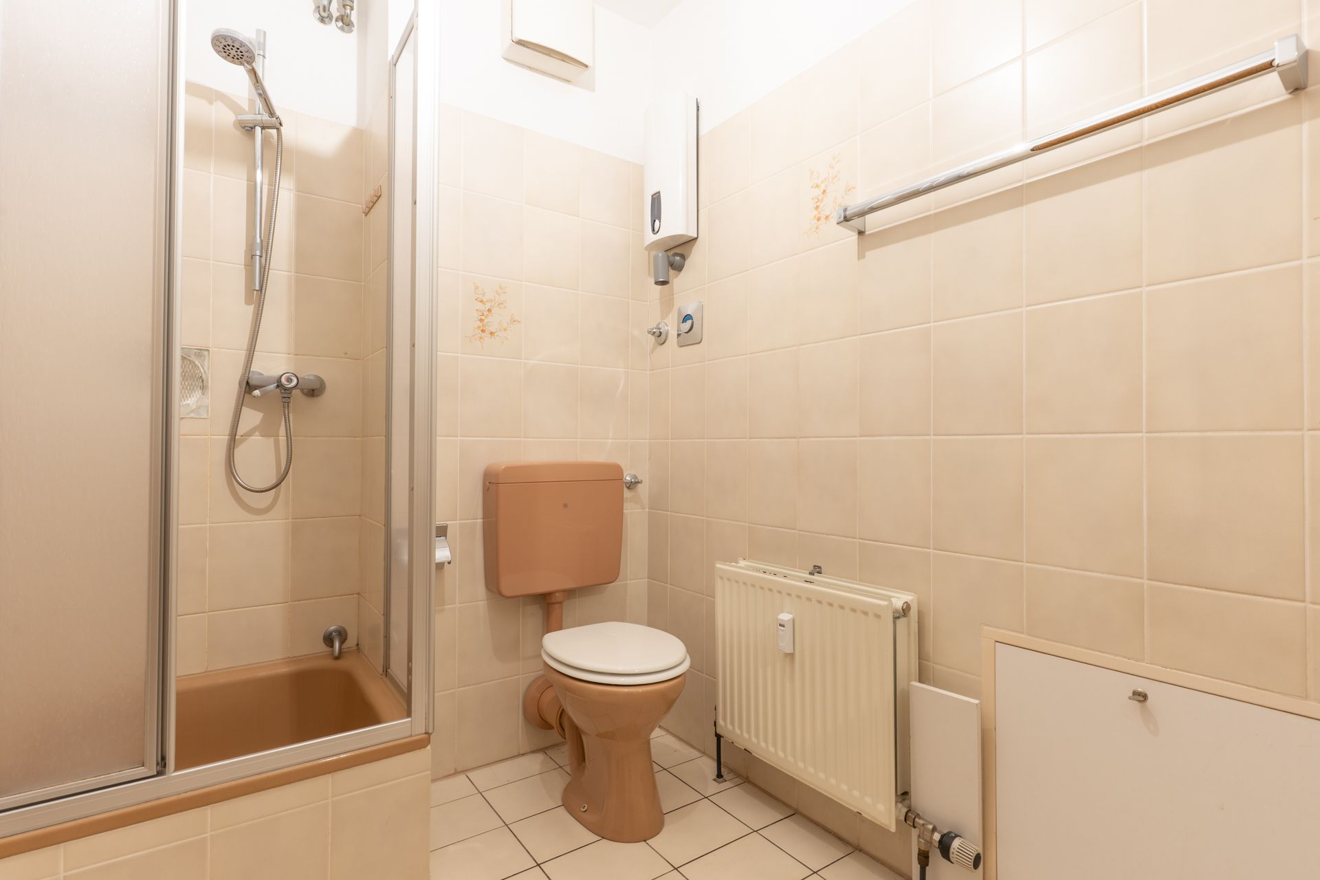 Kleines Badezimmer mit Dusche, Toilette und Heizkörper, beige Fliesen an Wand und Boden.