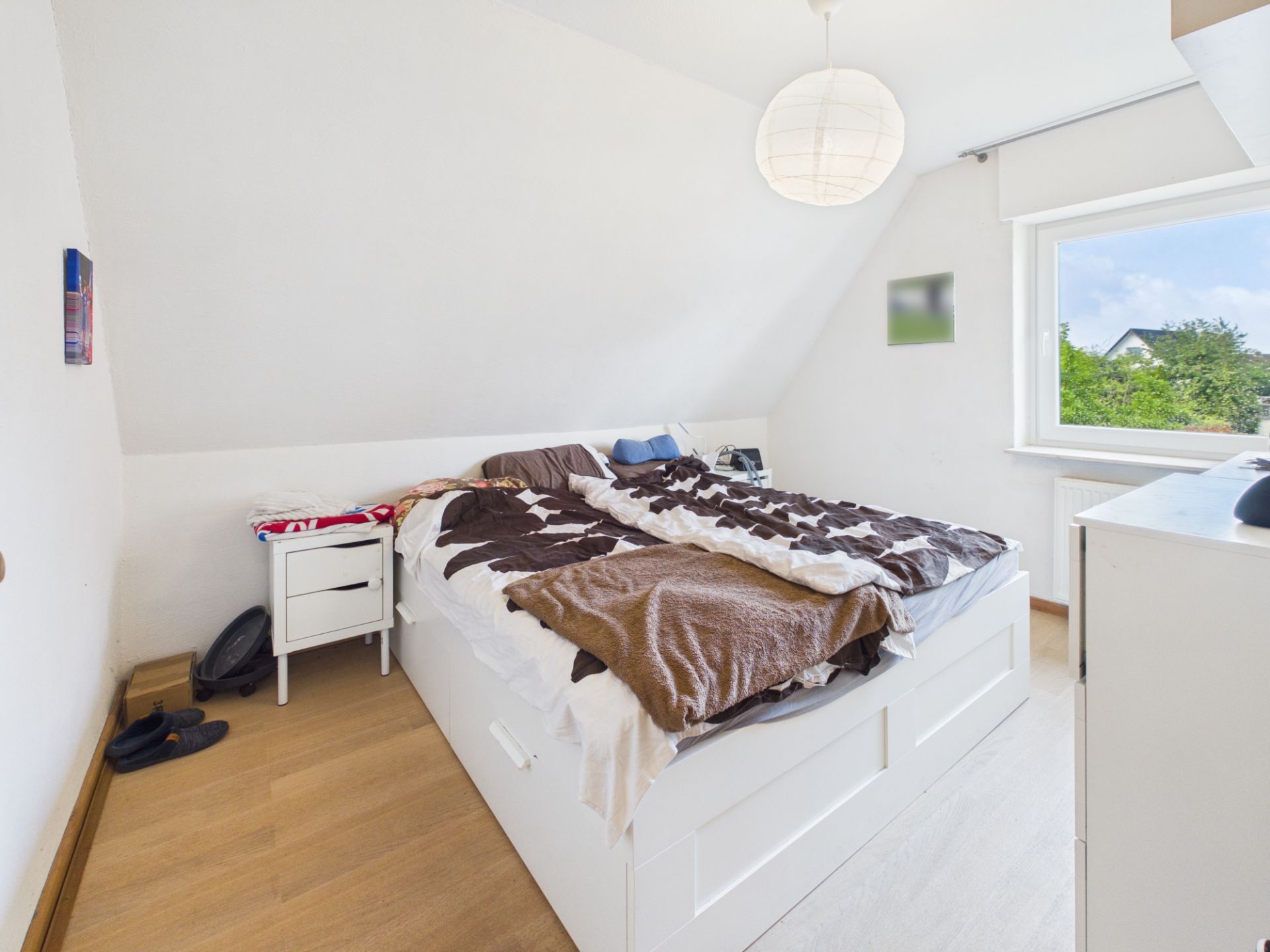 Helles Schlafzimmer mit Doppelbett, Nachttisch, Kommode und Fenster mit Blick ins Grüne.