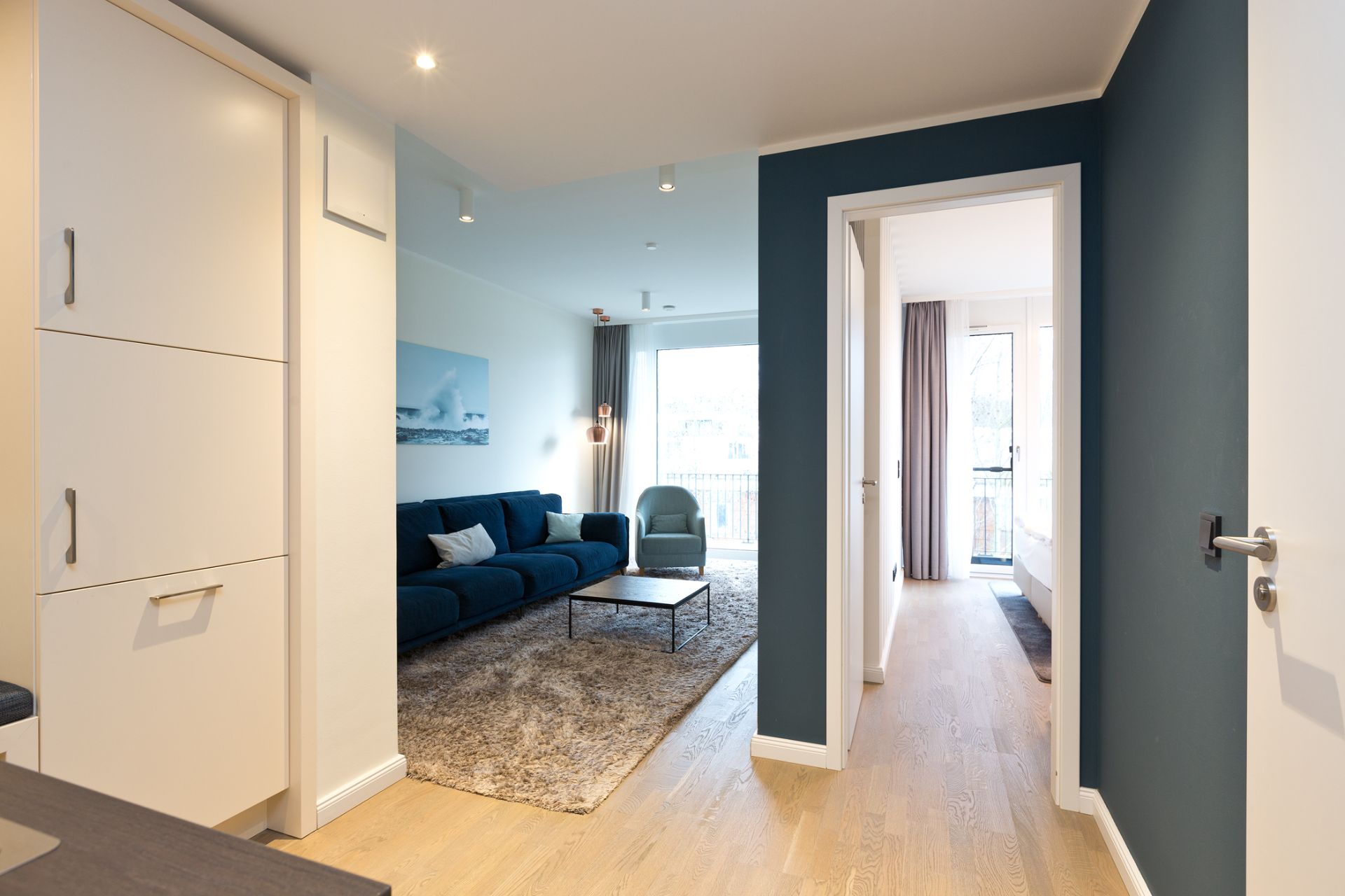 Helles Wohnzimmer mit blauem Sofa, Sessel, Teppich und Zugang zu einem weiteren Raum mit Fenster.