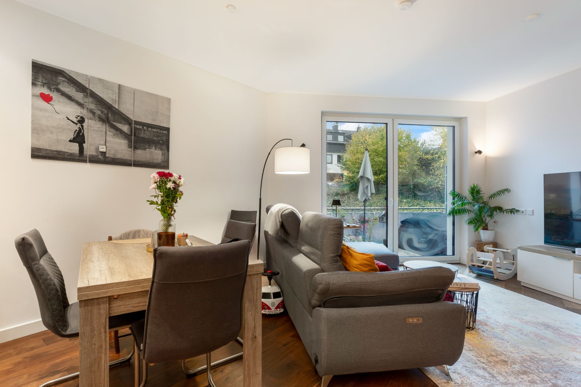 Wohnzimmer mit grauem Sofa, Esstisch, Stühlen und Zugang zu Balkon mit Blick ins Grüne.