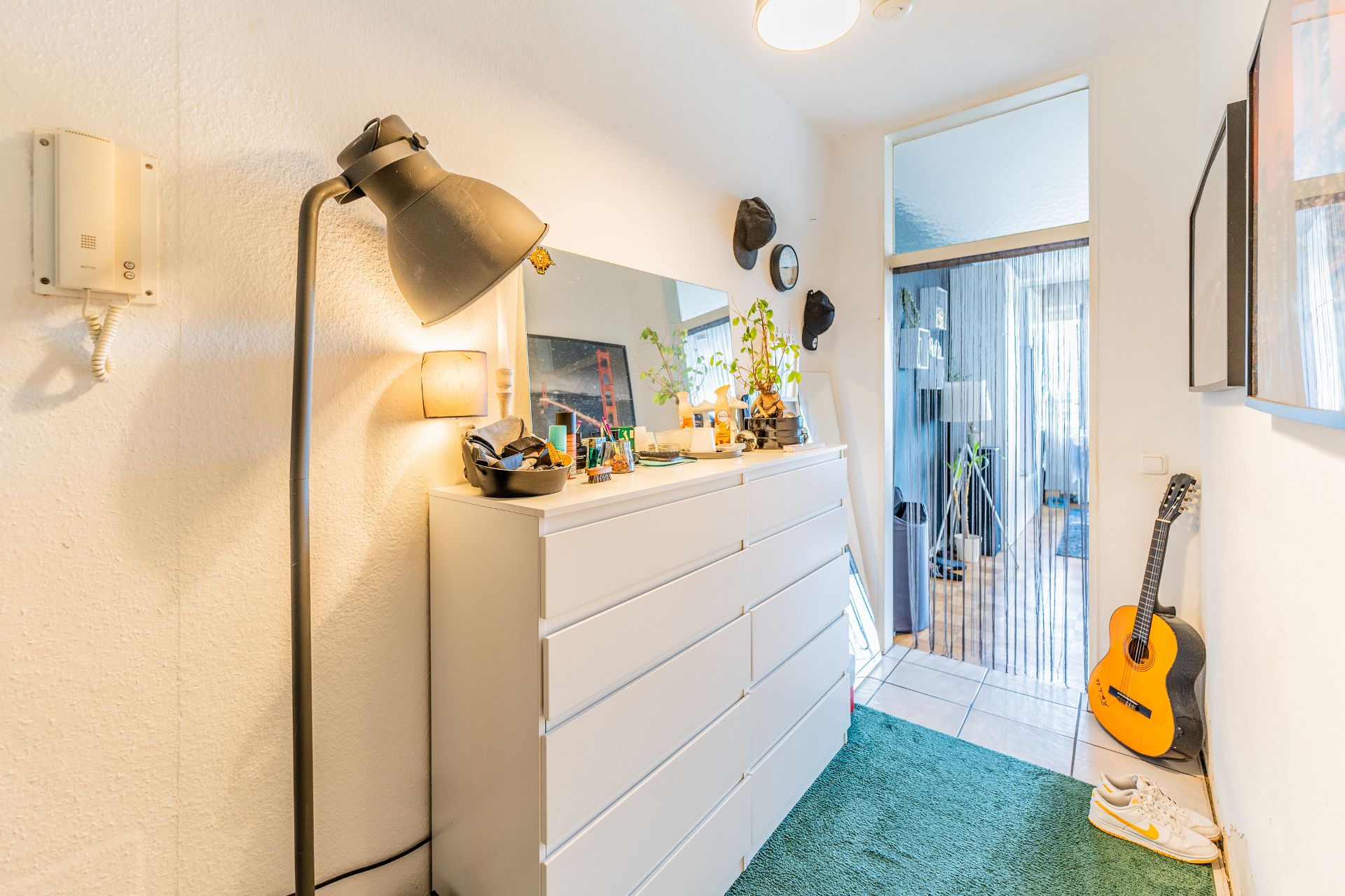 Heller Flur mit weißer Kommode, Spiegel, Stehlampe, Gitarre und grüner Teppichmatte.