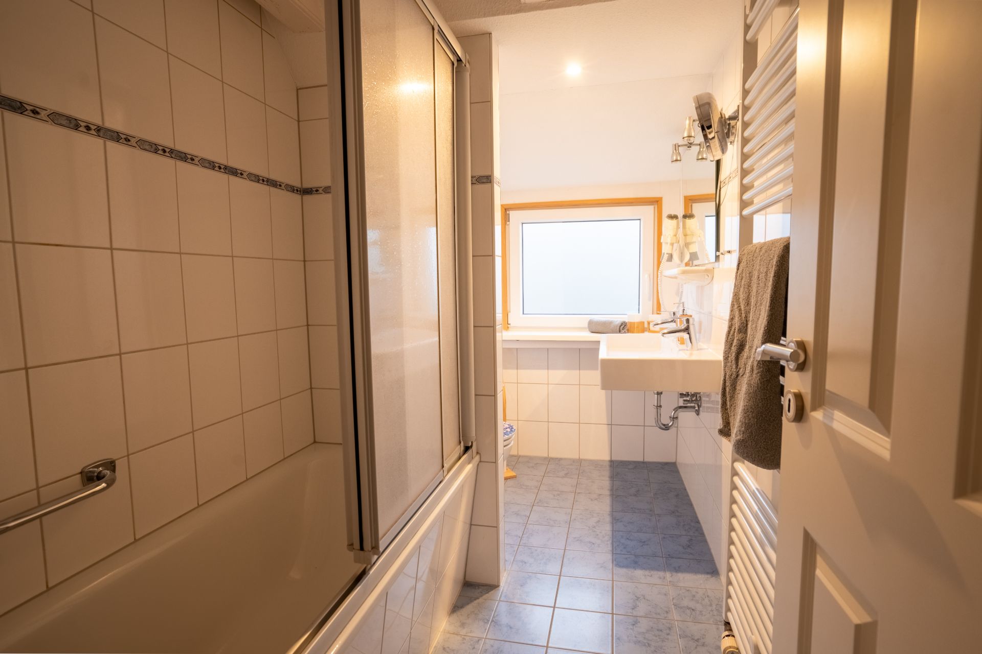 Helles Badezimmer mit Badewanne, Waschbecken, Fenster und Handtuch auf dem Heizkörper.