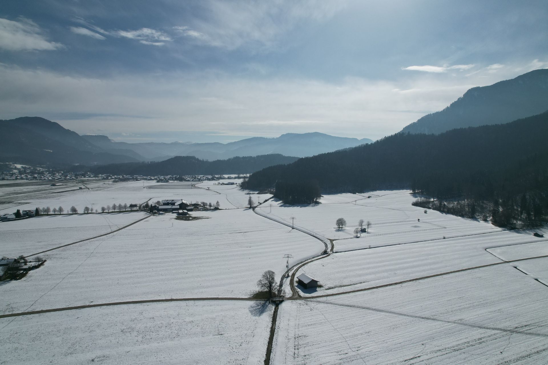 Schneebedeckte Felder mit einigen Bäumen und einem kleinen Haus, Berge im Hintergrund.