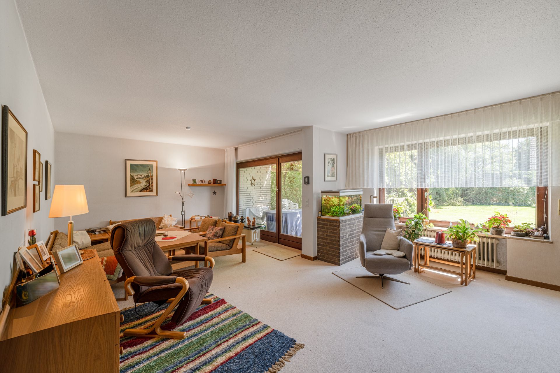 Helles Wohnzimmer mit Teppich, Sesseln, Esstisch, Aquarium und großen Fenstern mit Blick auf den Garten.