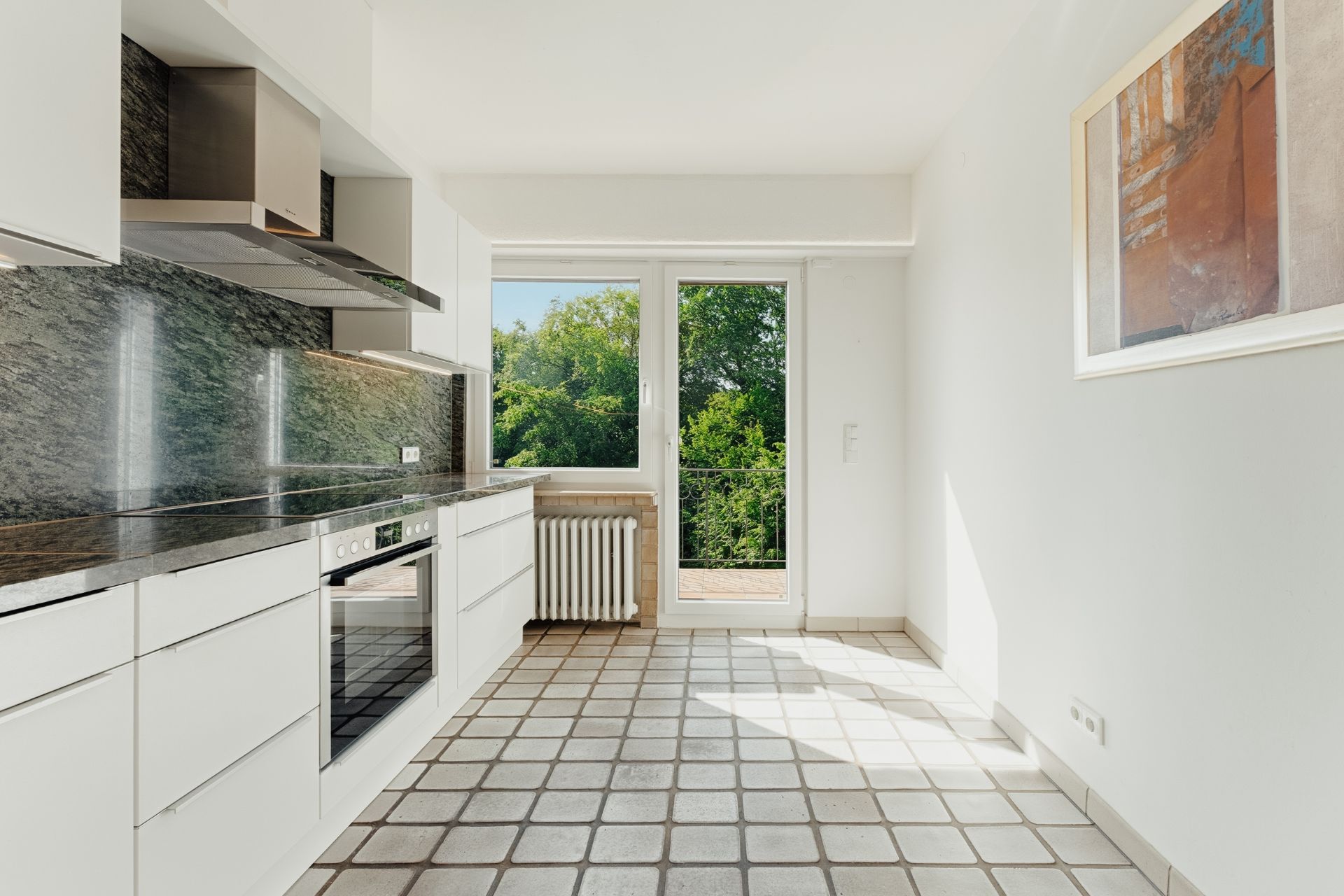 Helle Küche mit weißen Schränken, Fliesenboden, Fenster und Tür zu einem Balkon mit Blick ins Grüne.