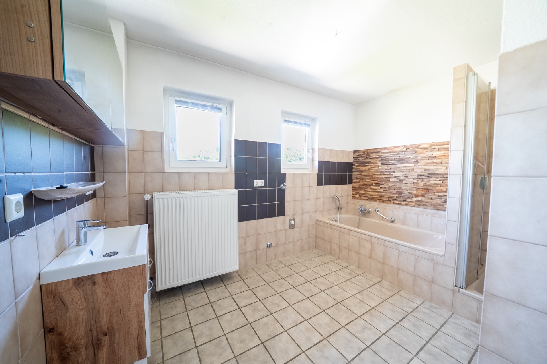 Helles Badezimmer mit Badewanne, Waschbecken, zwei Fenstern und gefliesten Wänden.