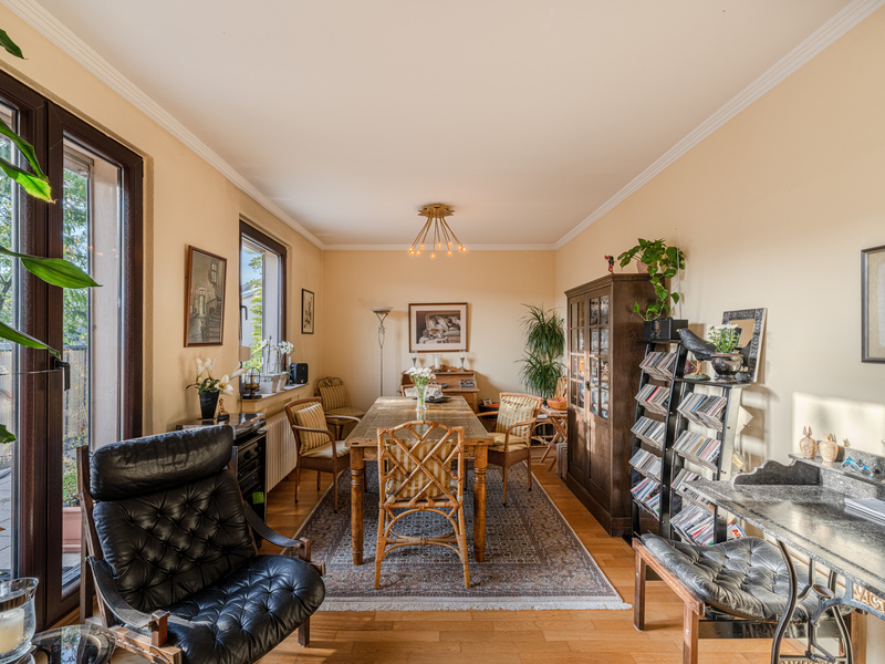 Helles Wohnzimmer mit Esstisch, Stühlen, Sesseln, Regal, Pflanzen und großen Fenstern.