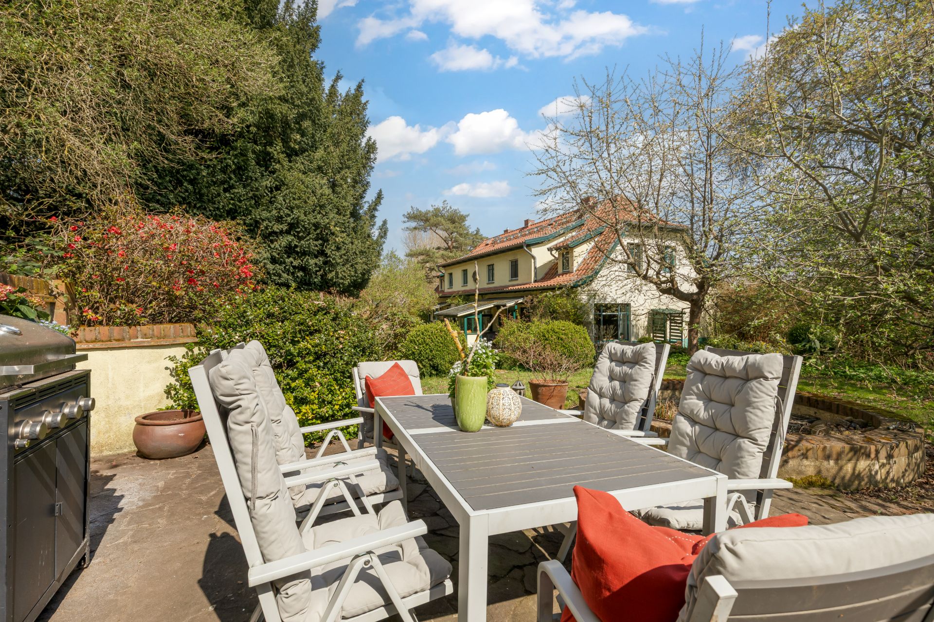Terrasse mit Gartentisch, sechs Stühlen und Grill, umgeben von Grünpflanzen, Haus im Hintergrund.