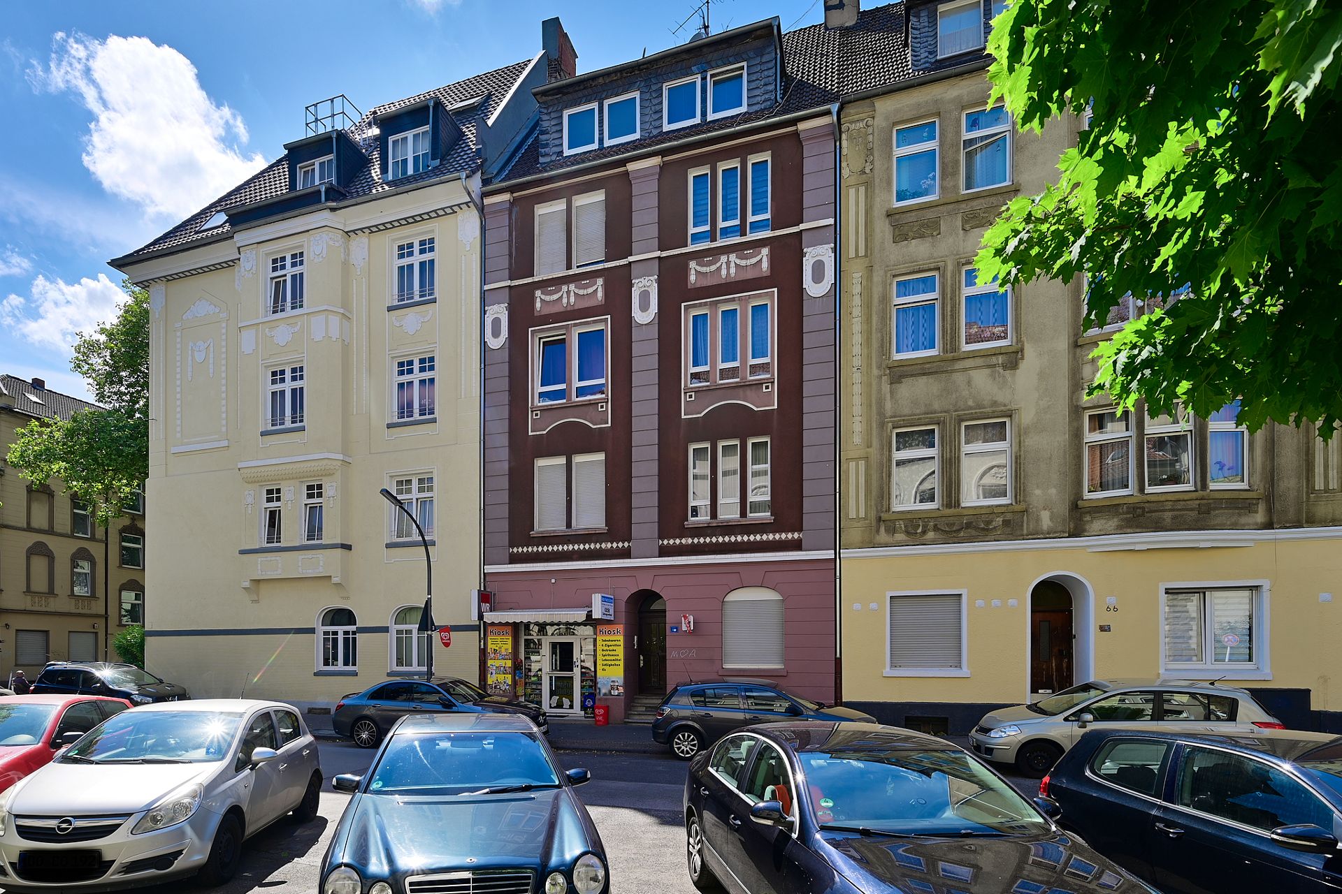 Mehrstöckige Wohnhäuser an einer Straße mit geparkten Autos, bei sonnigem Wetter.