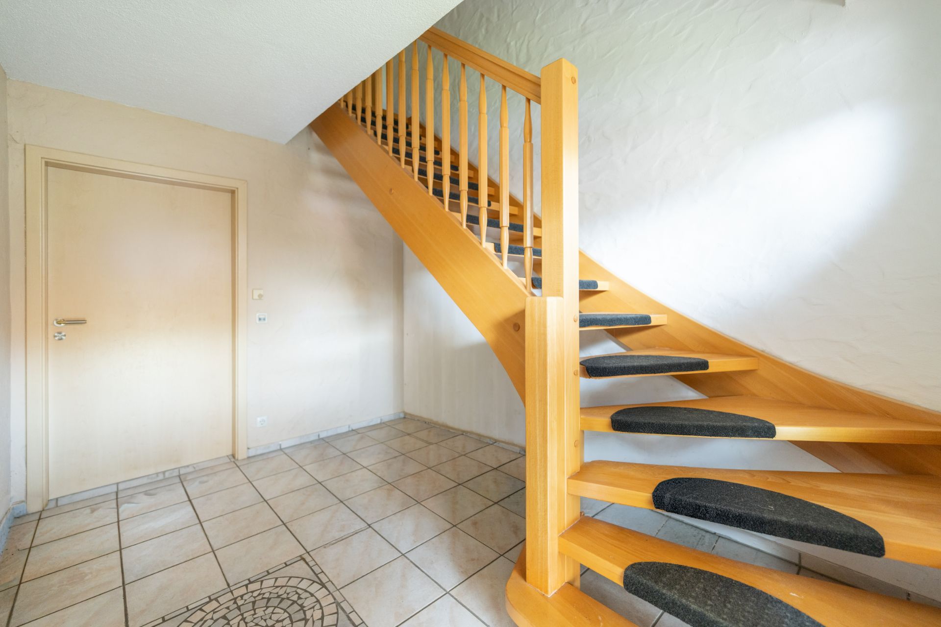 Holztreppe mit Teppichstufen in einem Flur mit Fliesenboden und heller Tür.