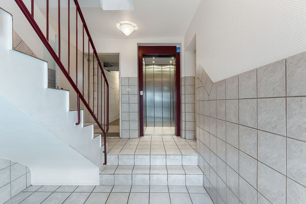 Treppenhaus mit Fliesenboden, rotem Geländer und Aufzug in einem Wohngebäude.