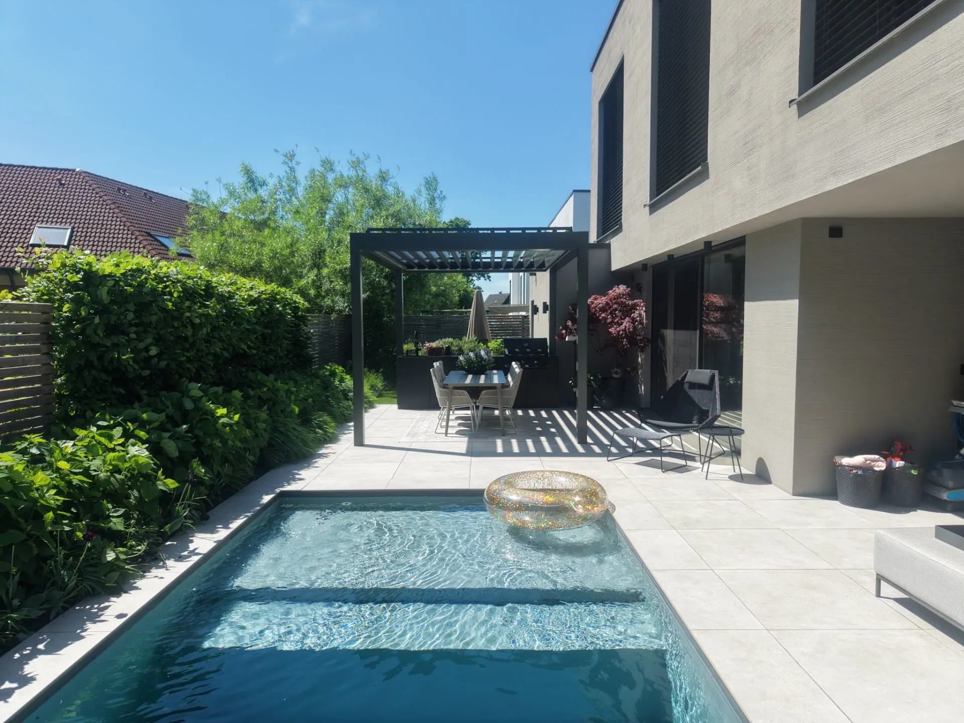 Moderne Terrasse mit Pool, Loungestühlen, Essbereich unter Pergola und grüner Hecke.