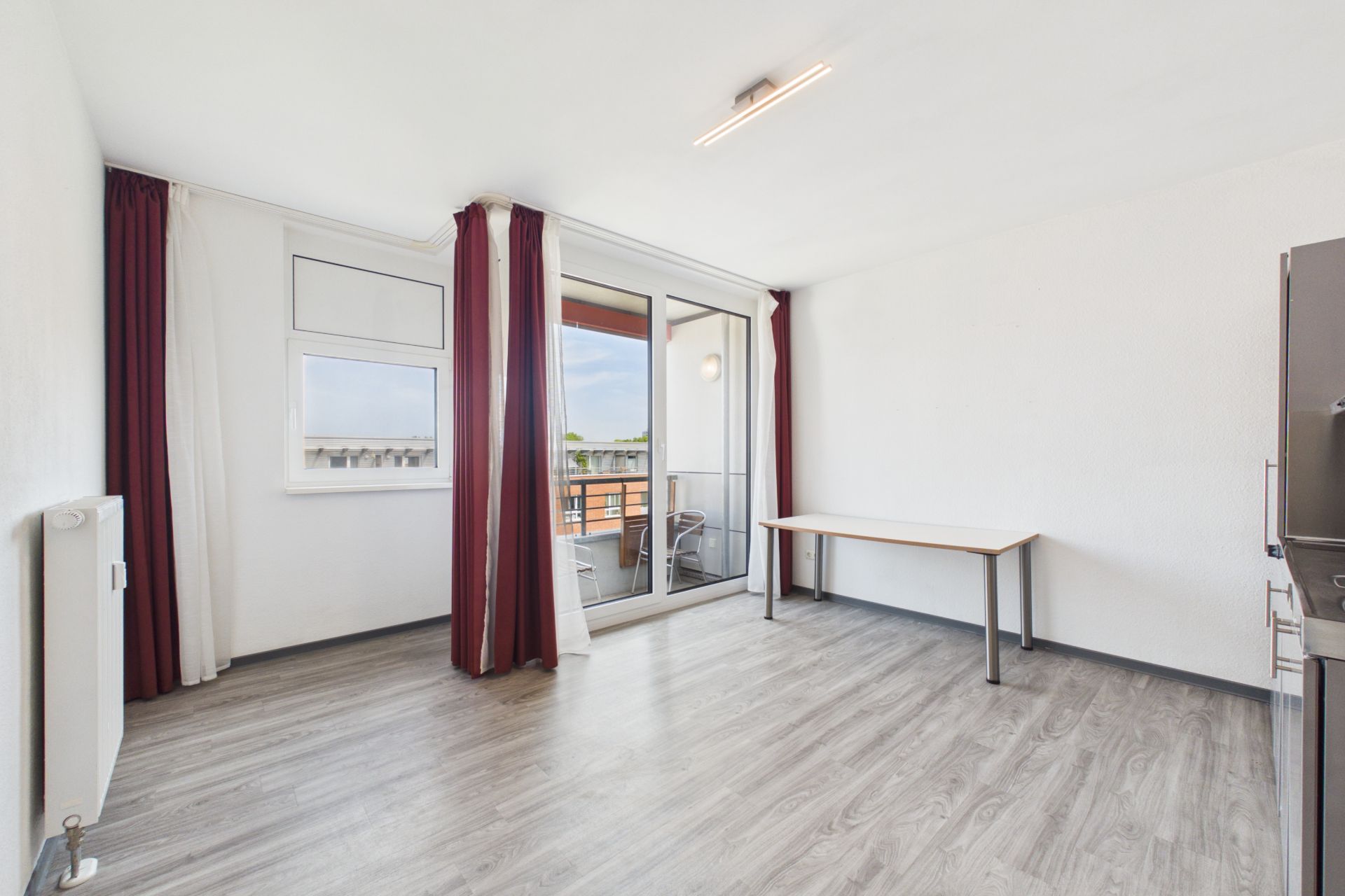Helles Zimmer mit Fenster, Balkonzugang, Tisch und Holzfußboden.