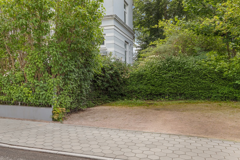 Unbefestigter Parkplatz neben Gehweg, umgeben von Büschen und einem weißen Haus.