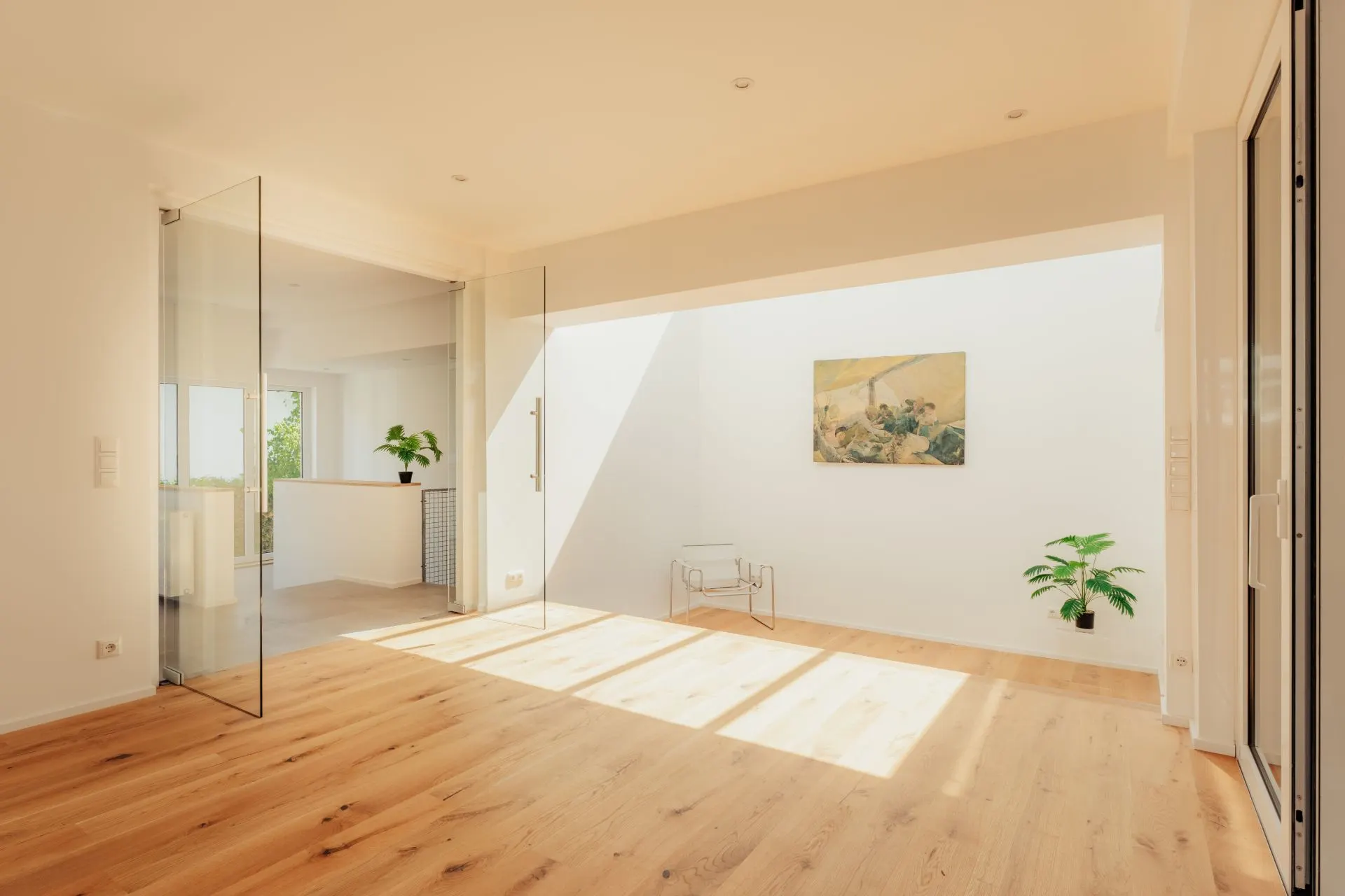 Heller Raum mit Holzboden, Glaswänden, zwei Pflanzen und einem Bild an der Wand.
