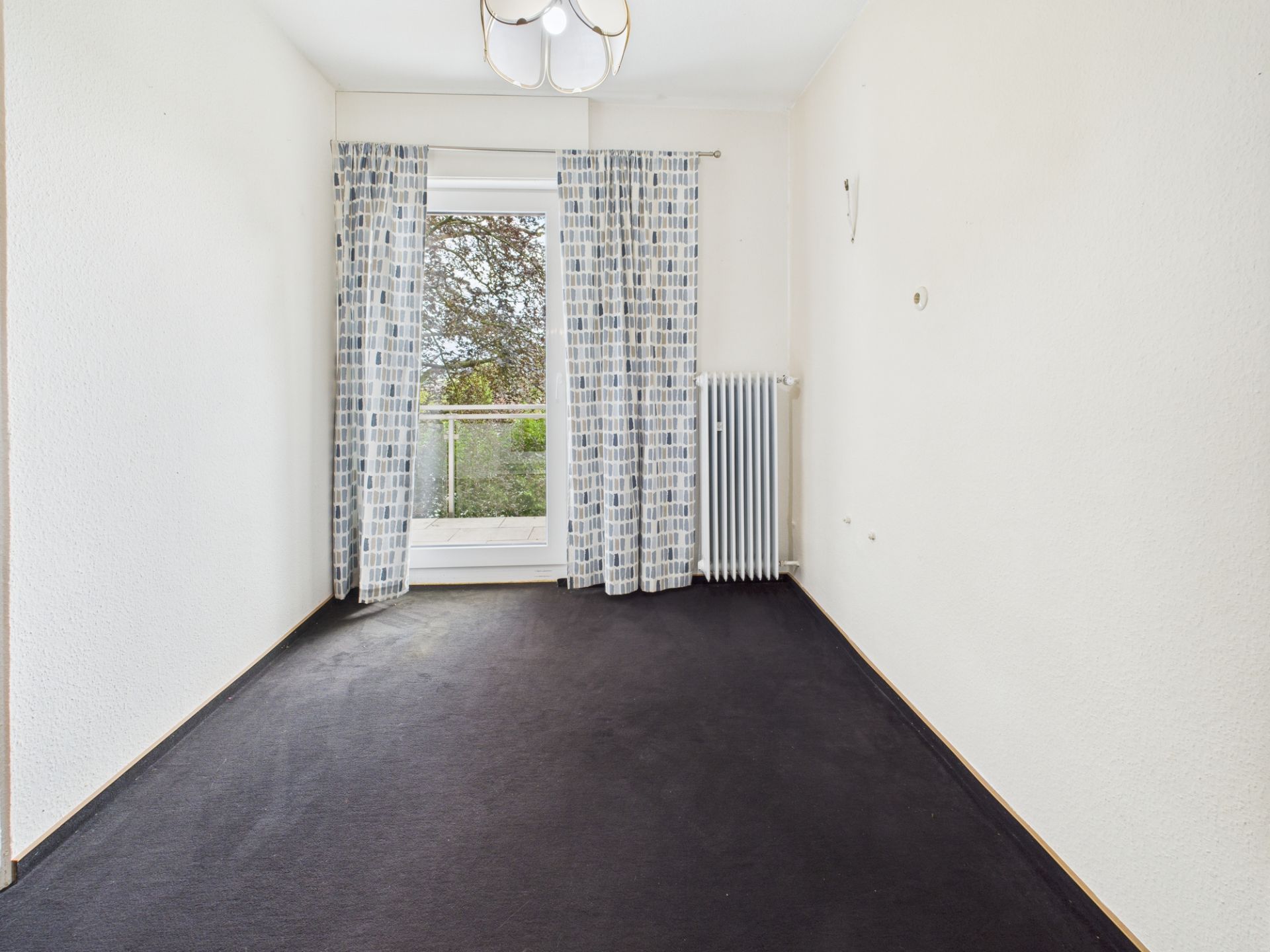 Leeres Zimmer mit dunklem Teppich, Vorhängen und Glastür zu einem Balkon.