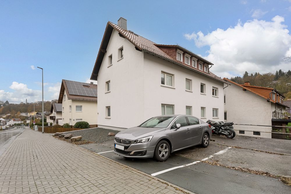 Weißes Wohnhaus mit schrägem Dach, davor geparktes Auto und Motorrad, gepflasterter Gehweg.
