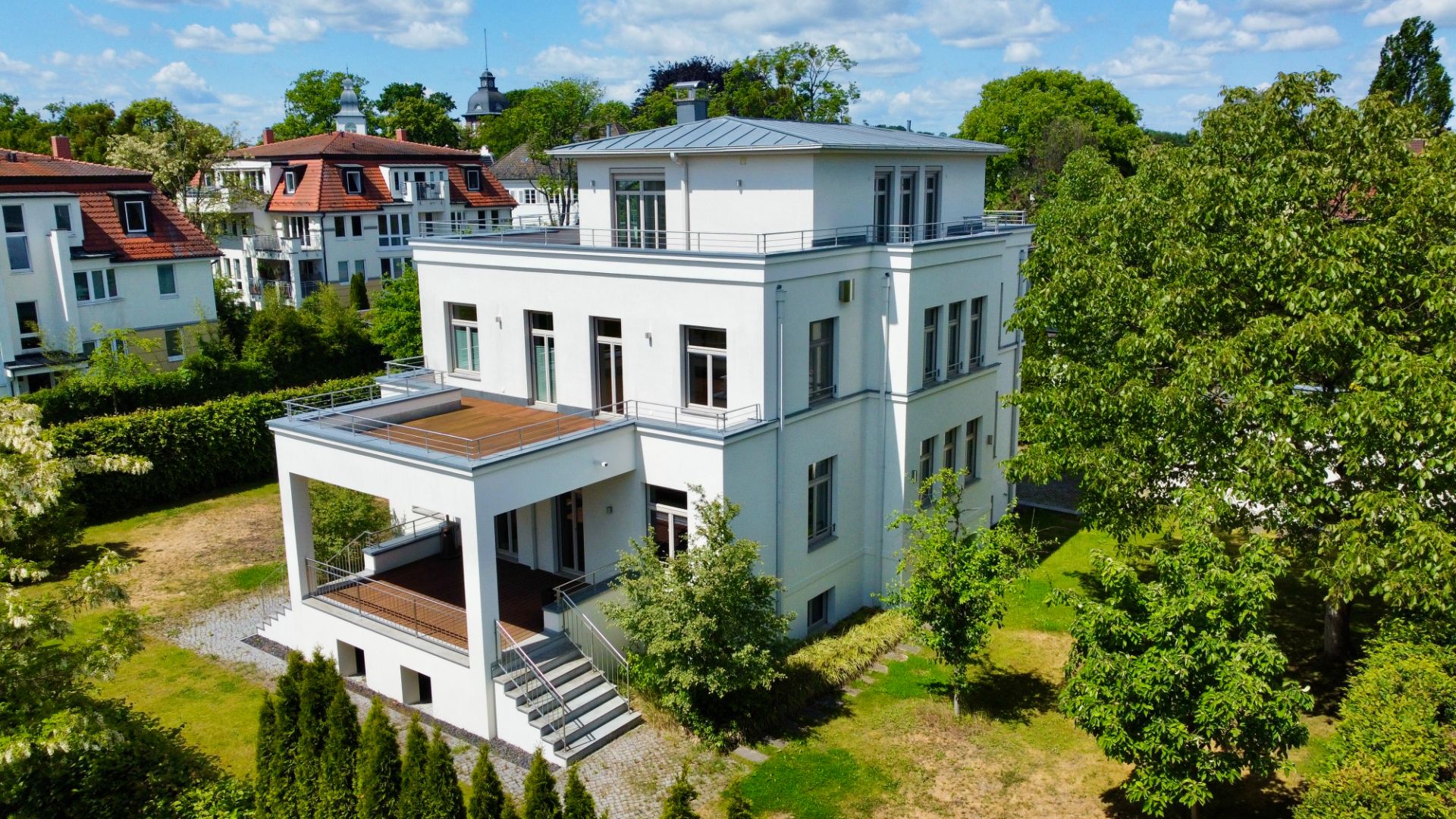 Modernes, weißes Wohnhaus mit mehreren Stockwerken, großer Terrasse und umgeben von Bäumen.