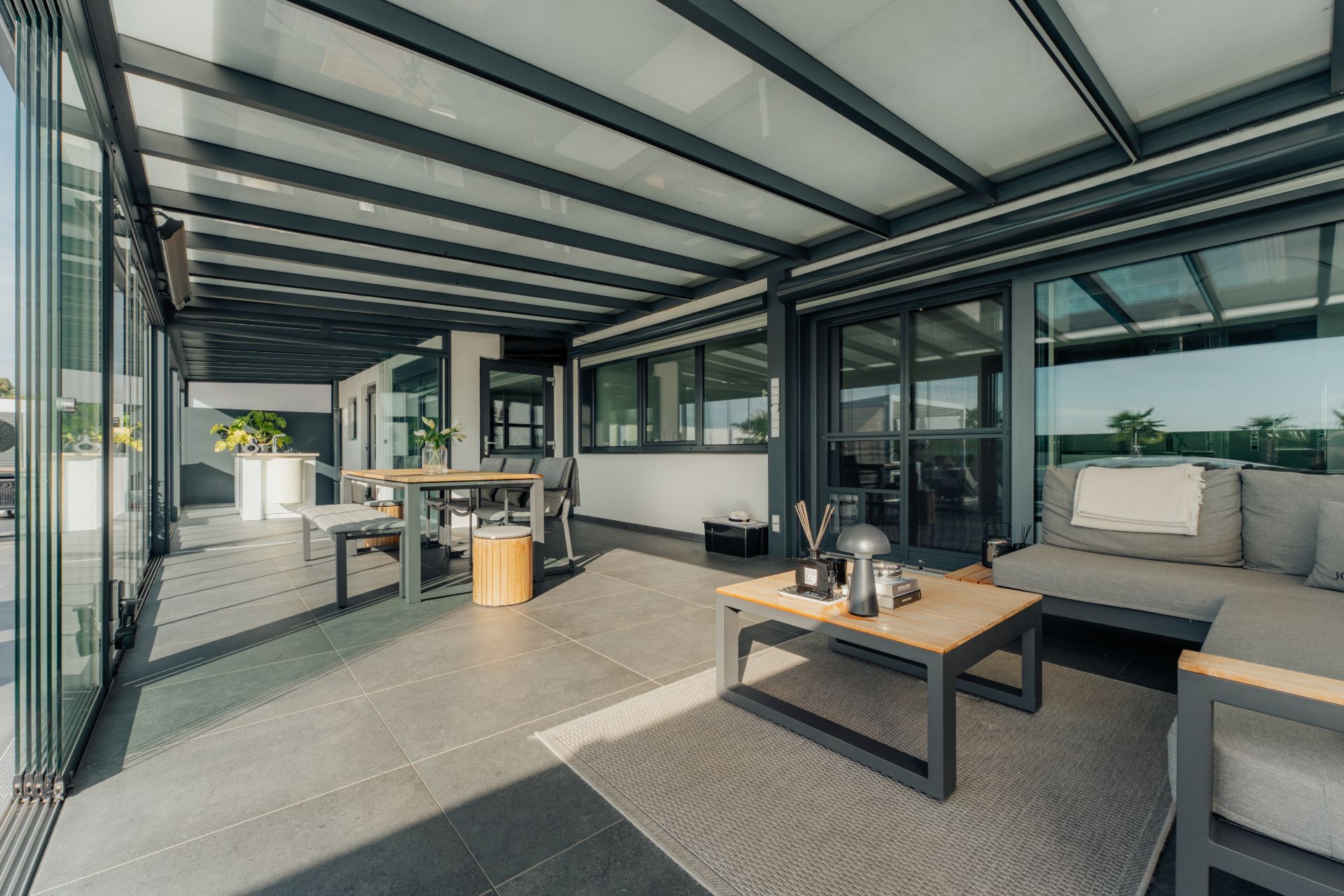Überdachte Terrasse mit Sitzbereich, Tisch und Sofa, große Fenster an der Seite.