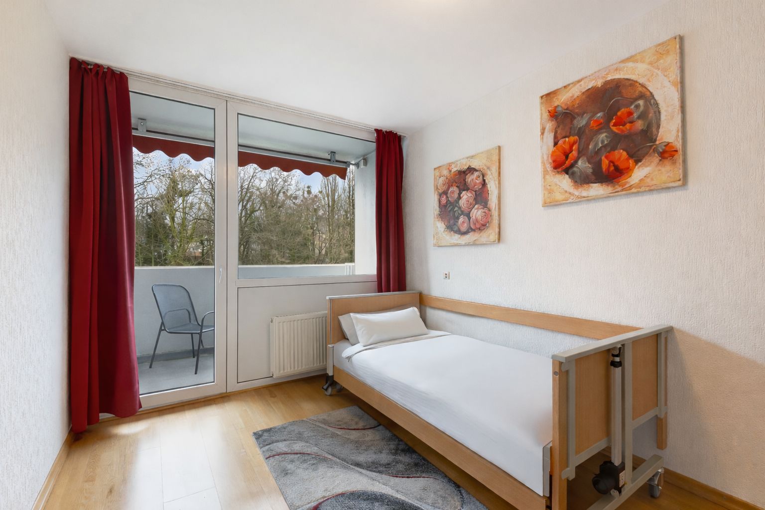 Ein Zimmer mit Einzelbett, hellen Wänden, rotem Vorhang und Zugang zu einem Balkon mit Stuhl.