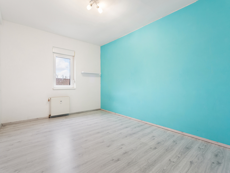 Leeres Zimmer mit hellem Boden, weißer Wand, türkisfarbener Wand und einem Fenster.