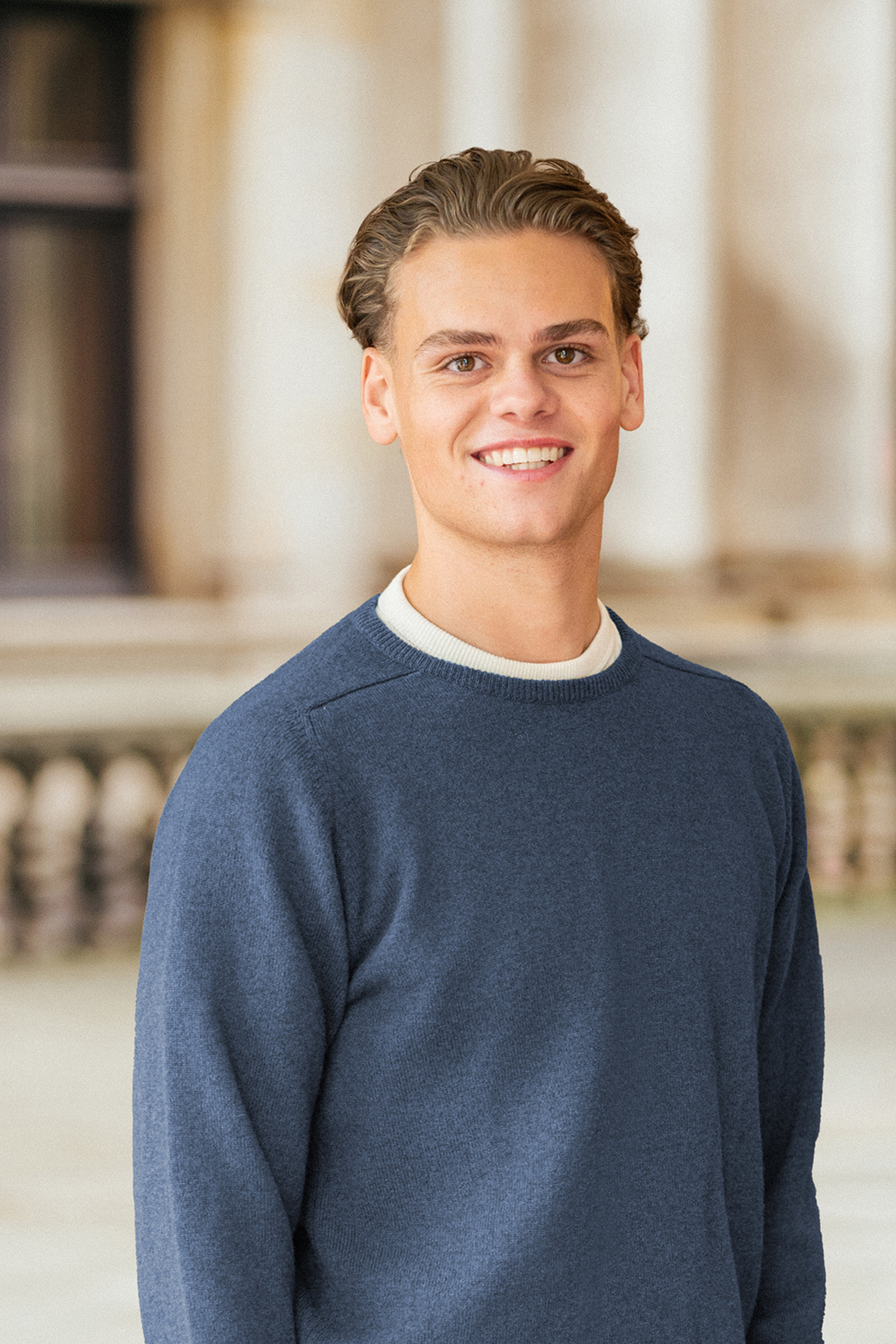 Junger Mann mit dunkelblauem Pullover steht vor einem Gebäude und lächelt.