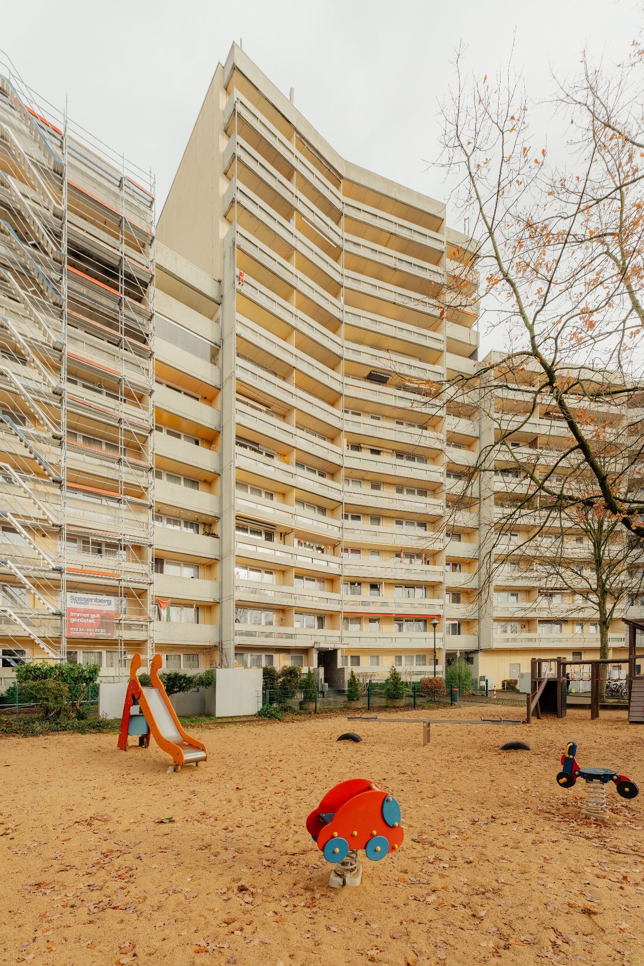 Mehrstöckiges Wohnhaus mit Balkonen, davor ein Sandspielplatz mit Rutsche und Wippe.