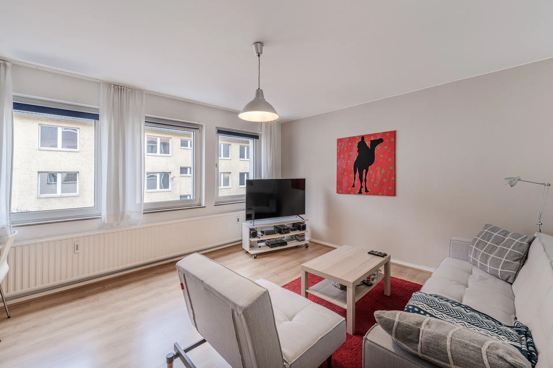 Helles Wohnzimmer mit Sofa, Sessel, Couchtisch, Fernseher und drei großen Fenstern.