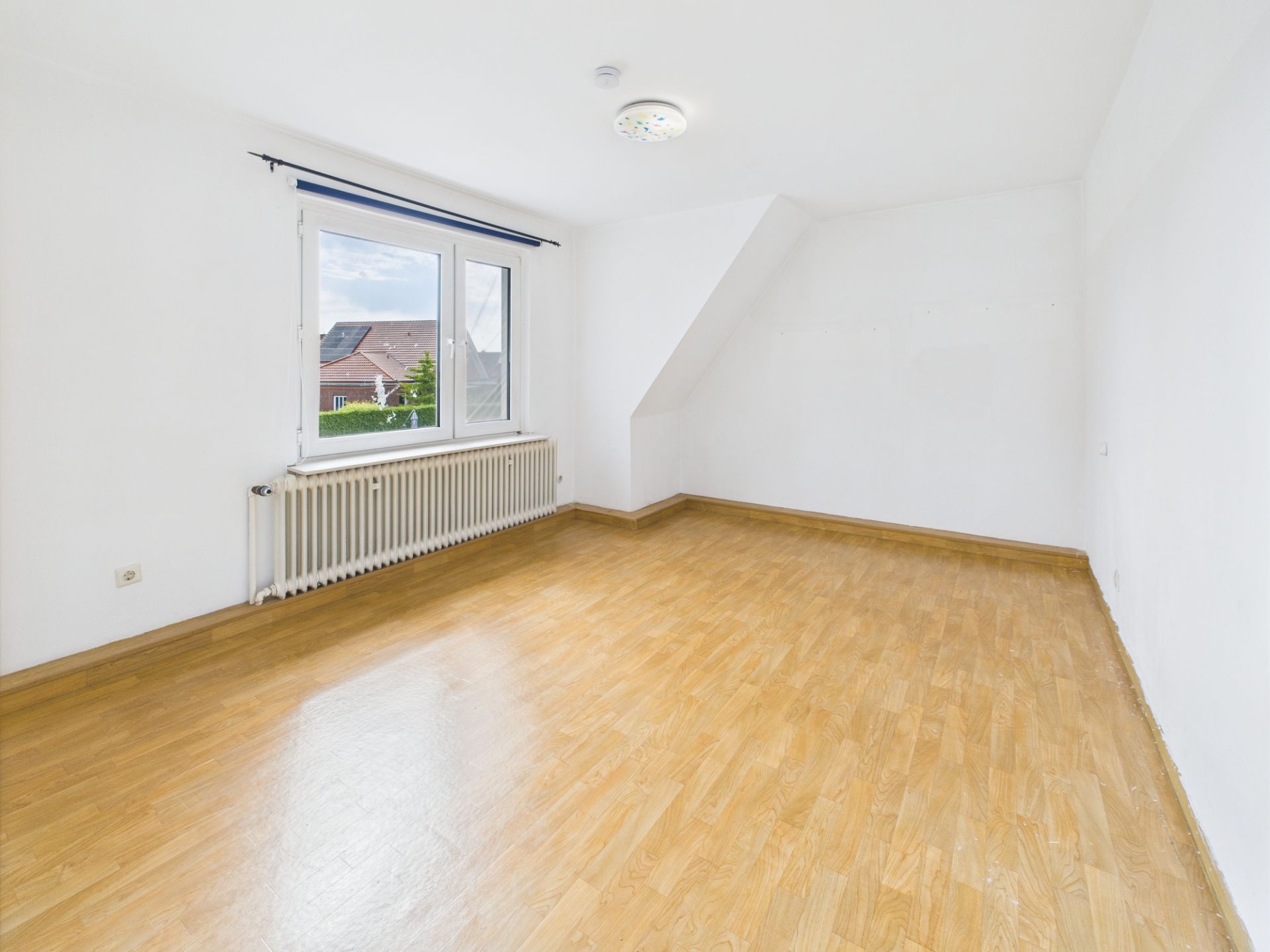 Leerer Raum mit Holzfußboden, weißer Wand und großem Fenster mit Heizung darunter.