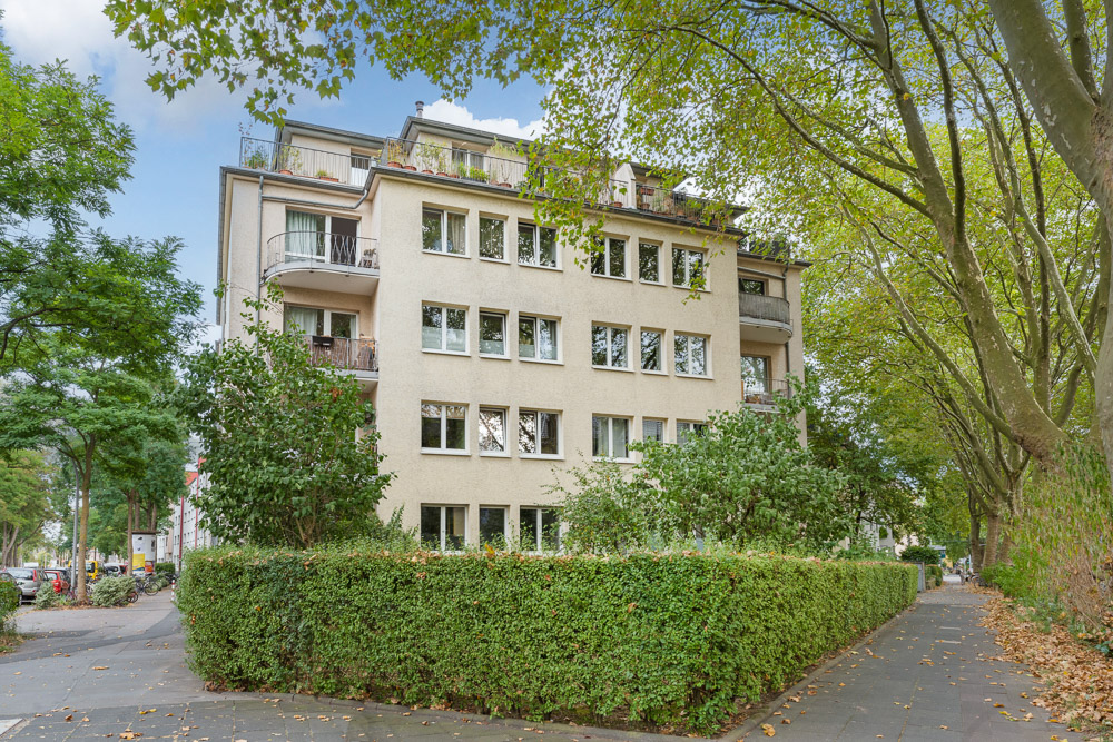 Mehrstöckiges Wohnhaus mit Balkonen, umgeben von Bäumen und Hecken an einer Straßenecke.