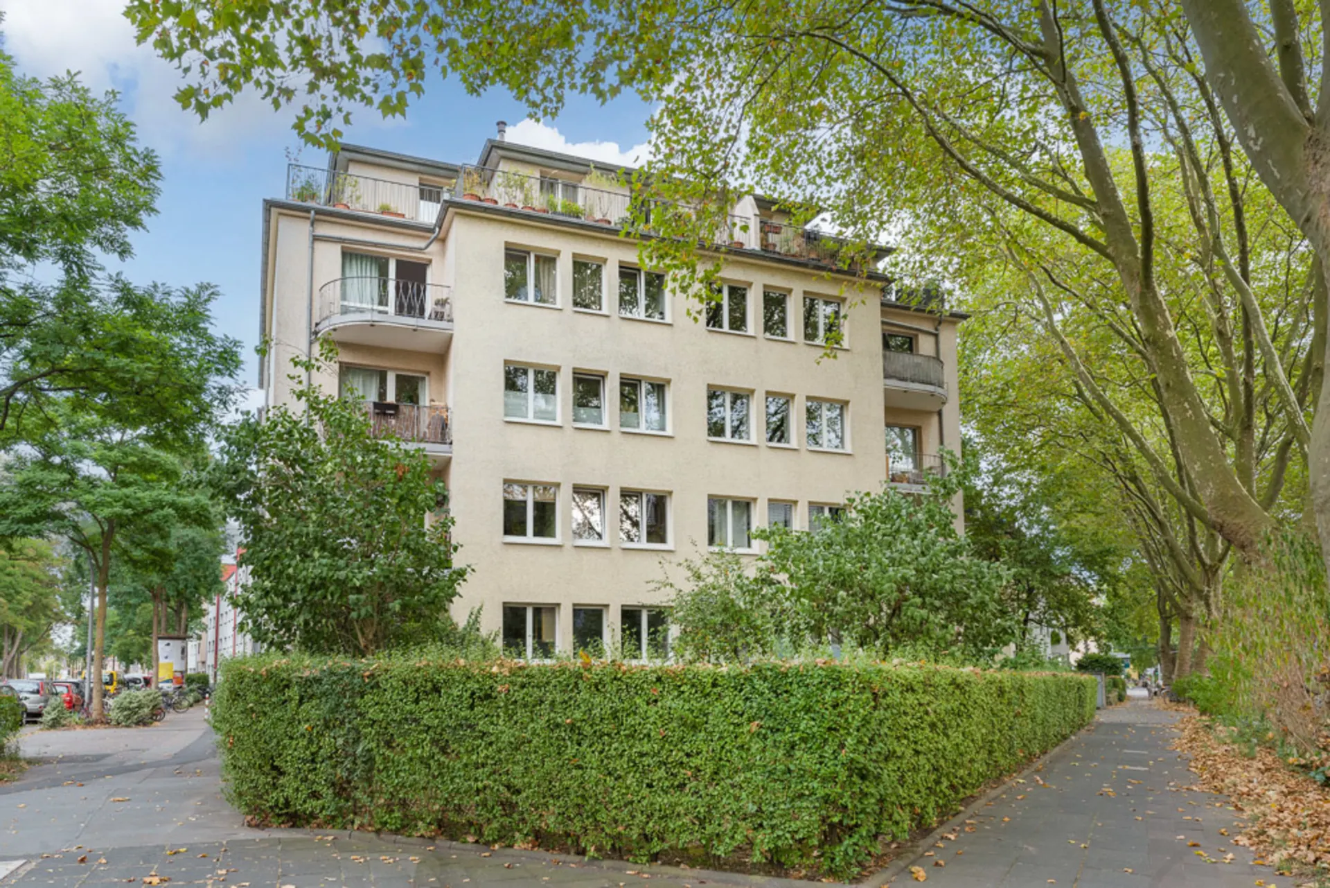 Mehrstöckiges Wohnhaus mit Balkonen, umgeben von Bäumen und Hecken an einer Straßenecke.