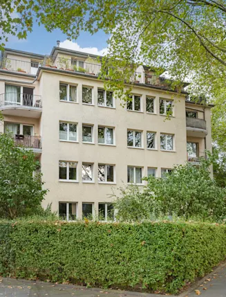 Mehrstöckiges Wohnhaus mit Balkonen, umgeben von Bäumen und Hecken an einer Straßenecke.