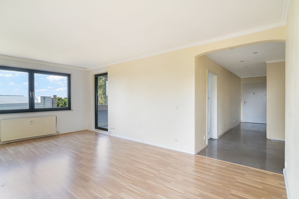 Helles Wohnzimmer mit Holzboden, großem Fenster, Tür zum Balkon und Flur mit Fliesenboden.