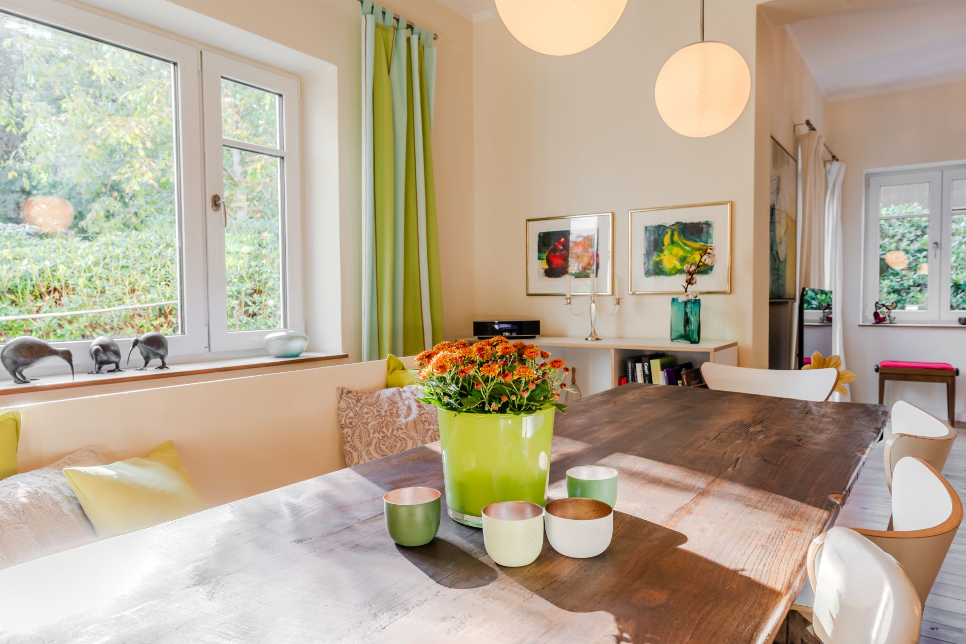 Helles Esszimmer mit Holztisch, grüner Pflanze, Fenstern und Wandbildern.