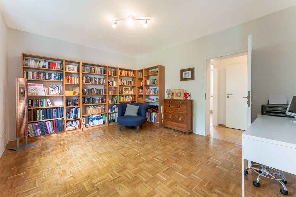 Helles Zimmer mit Bücherregalen, Sessel, Kommode und Schreibtisch auf Parkettboden.