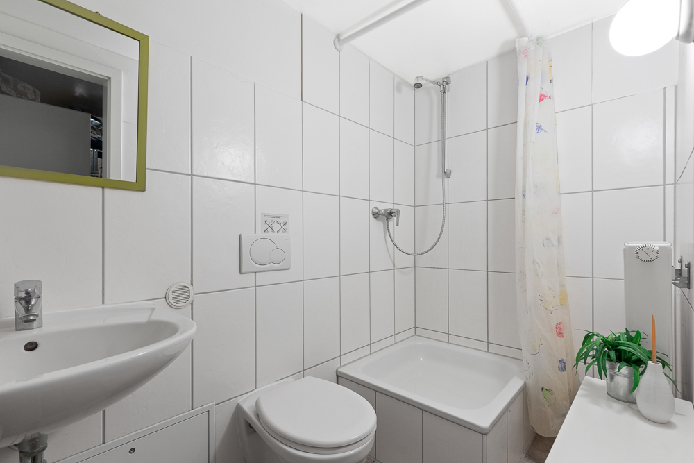 Kleines Badezimmer mit Dusche, WC, Waschbecken, Spiegel und Pflanze auf weiß gefliesten Wänden.