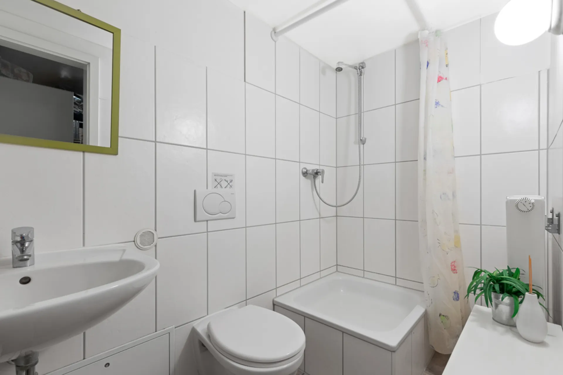 Kleines Badezimmer mit Dusche, WC, Waschbecken, Spiegel und Pflanze auf weiß gefliesten Wänden.