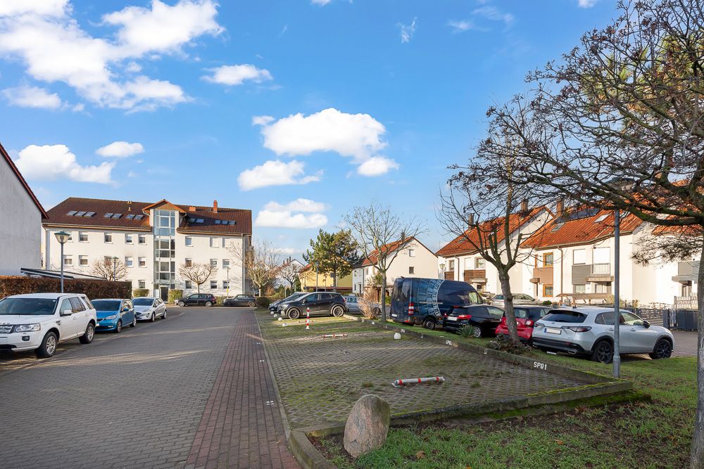 Wohnstraße mit parkenden Autos, Bäumen und Mehrfamilienhäusern bei blauem Himmel.
