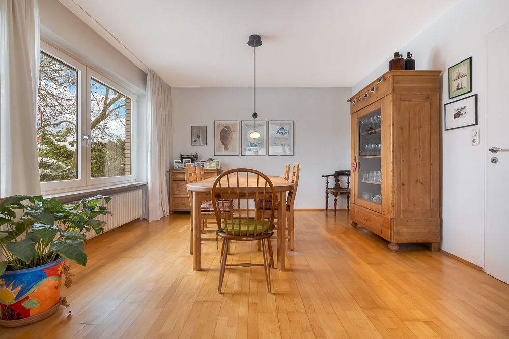 Esszimmer mit Holzboden, Holztisch, Stühlen, Vitrine, Pflanzen und großem Fenster.