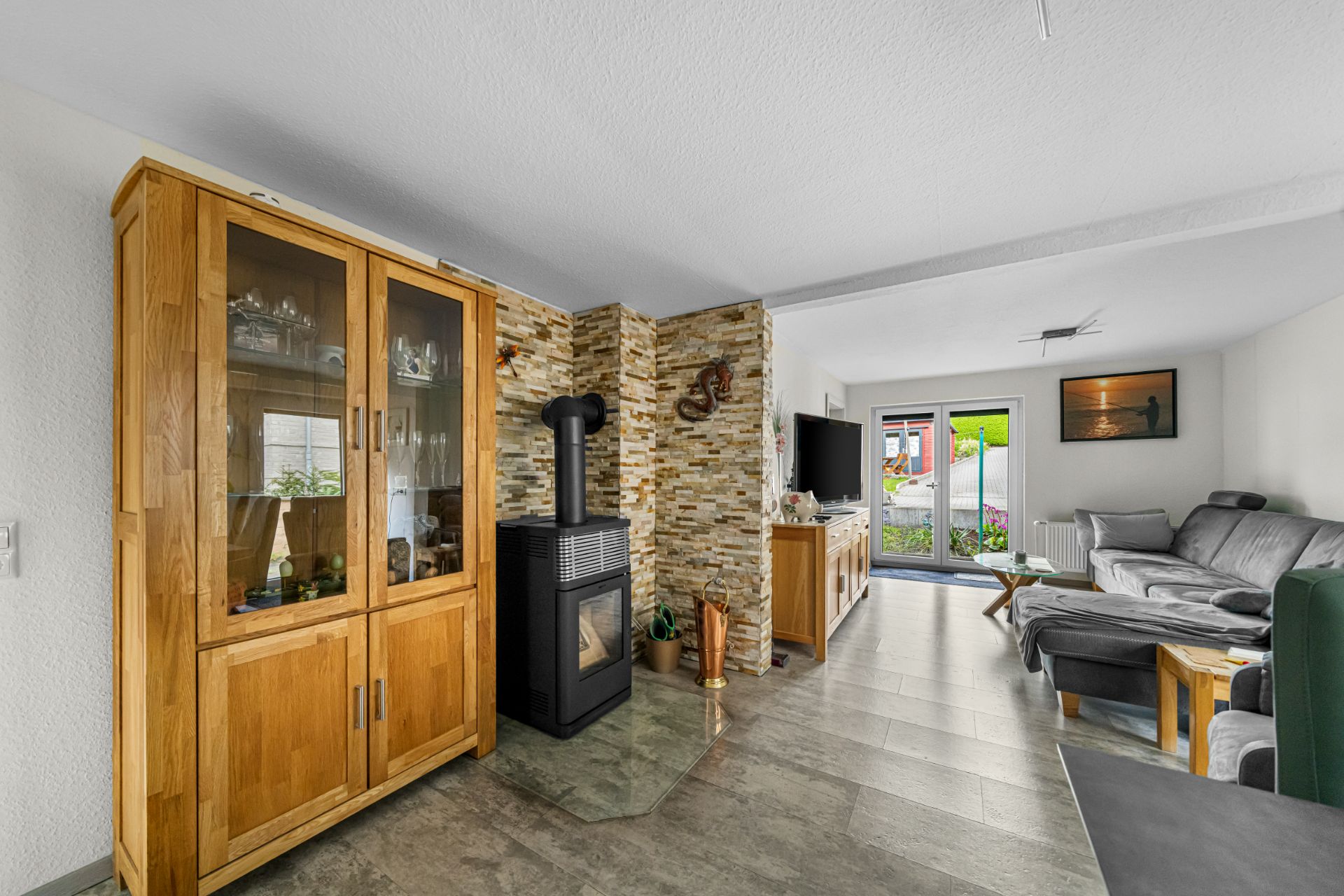 Wohnzimmer mit Holzschrank, Kaminofen, Sofa und Zugang zur Terrasse.